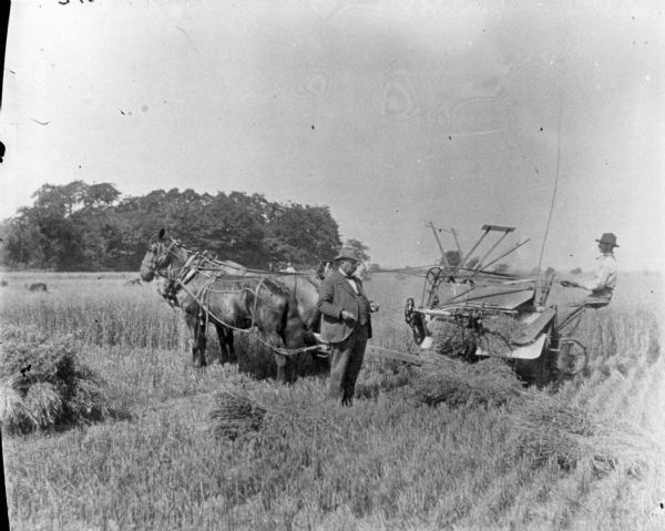 A man in a suit and hat is standing in front of a man using a horse-drawn binder in a field.