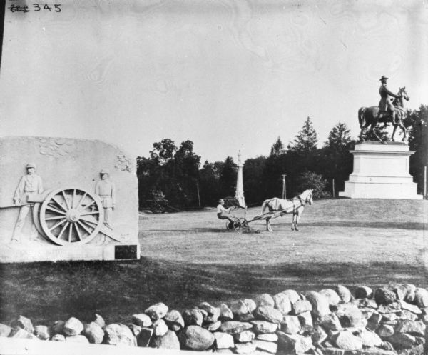 View over stone wall towards a man using a horse-drawn mower among various monuments and military cannons. The monument on the left is for "Rickett's Battery."