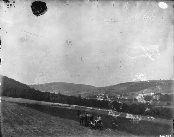Landscape view down hill towards man sitting on a mule-drawn binder in a field. In the background is a valley.