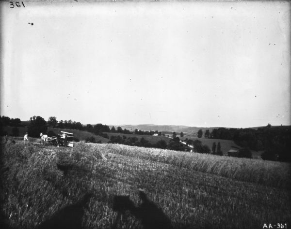 Landscape view across field with sheaves of wheat towards two men standing near a horse-drawn binder. A valley with buildings and trees is in the background. In the foreground are shadows of two men.