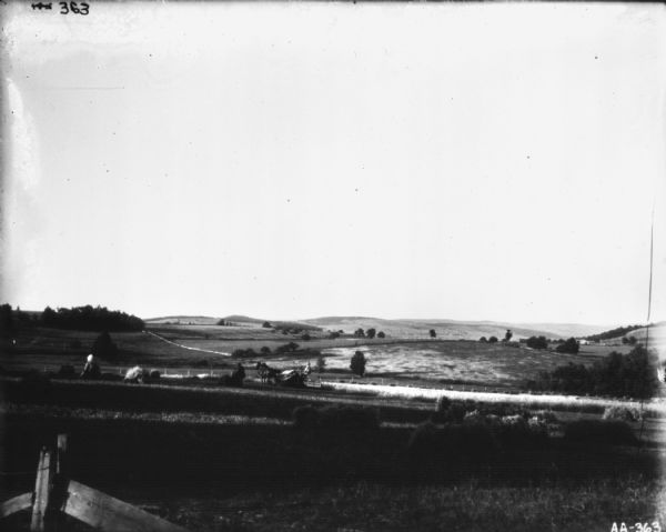Landscape view across field towards a man using a horse-drawn mower in a field. Other men are working in the field with sheaves of wheat. There is a fence in the foreground.