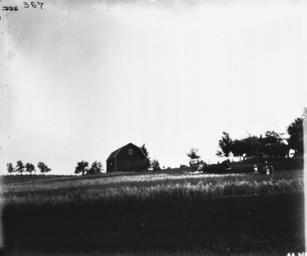 View across field towards a woman standing with two children. In the field a man is using a horse-drawn binder. A barn and farmhouse are in the background.