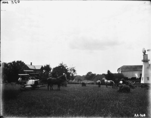 Right side view of a man using a horse-drawn binder. Another man is standing in the field next to two people sitting in a horse-drawn carriage. On the right is a windmill and a barn. A farmhouse is in the background on the left.