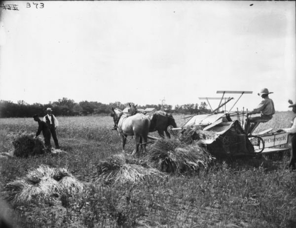 Three-quarter view from left rear of a man using a horse-drawn binder in a field. Two men are standing near a sheaf of wheat on the left. Another man is standing behind the binder on the far right. Buildings are in the far background.
