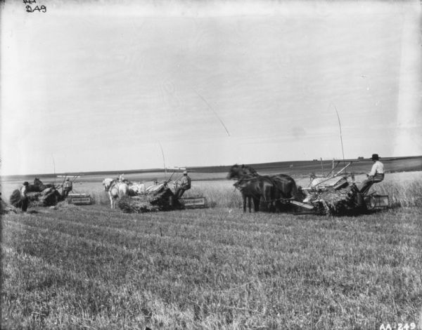 Slightly elevated view of six men and three children in a field. In the center a man is using a horse-drawn mower. On the left a man is sitting in a horse-drawn carriage, and two men are standing near the horse. In the center a man is standing with three children, and on the right another man is standing in front of a horse-drawn carriage. In the background in the field are piles of hay.