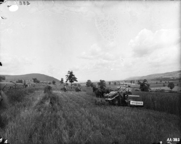 Landscape view of a man using a horse-drawn binder in a field surrounded by a fence. Two men in the background near the fence are collecting sheaves of hay. In the far background are more fields in a valley.