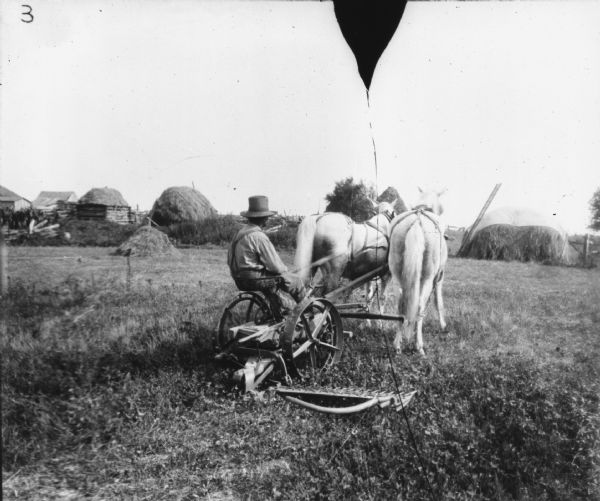 Rear view of a man using a horse-drawn mower in a field. In the background are large stacks of hay near fences and farm buildings.