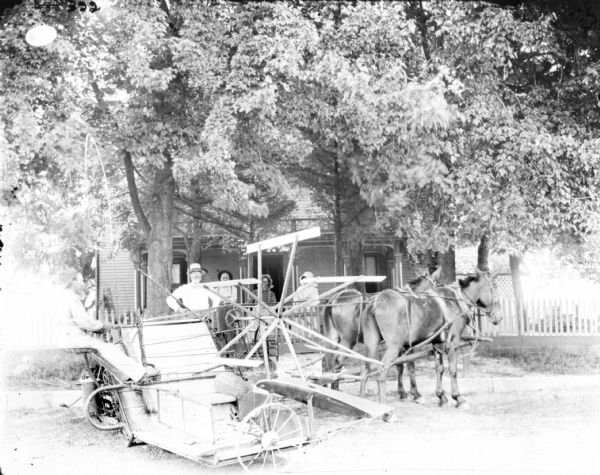 A man is sitting on a mule-drawn binder in the foreground. In the background two men, and two women wearing bonnets, are standing on the porch of a house which is under a stand of trees.