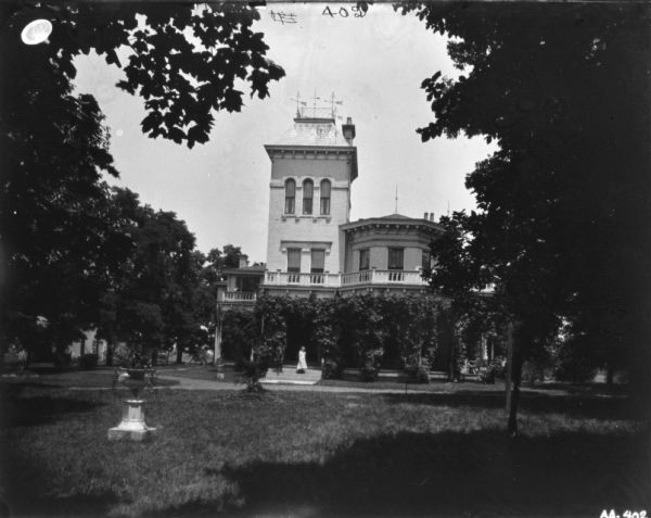 View across lawn towards large brick house, with wrap-around porch and balcony. A young boy and girl are standing on the porch steps near the walk. On the top of the tower are four windvanes.