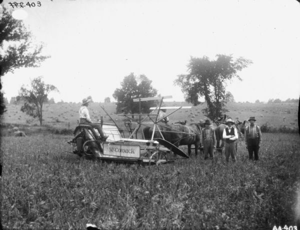 View across field of a group of three men standing near a man on horse-drawn binder. Another man is sitting on one of four horses pulling the binder.