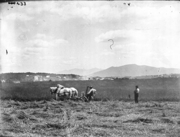 Man using a team of horses to pull a mower in a field. Another man is standing and watching on the right. There are buildings on a hill in the distance on the left, and a mountain in the far right background.