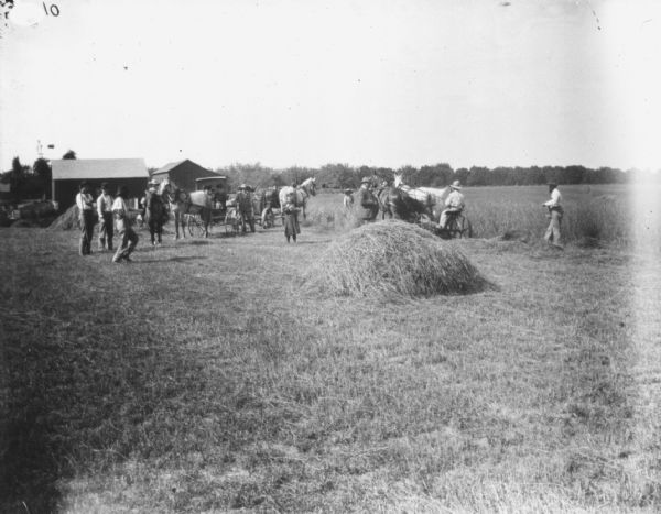 People are gathered in a field near a man using a horse-drawn mower in a field. There are horse-drawn buggies and farm buildings on the left.