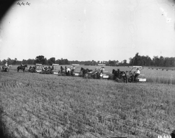 Slightly elevated view of a large group of people posing in a field. There are five men on five McCormick binders. A child is standing in front of one of the binders. Other men are standing among the binders, and another two men are on horseback on the left.