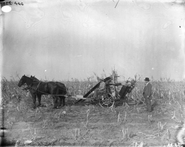Left side view of a man using a horse-drawn corn binder in a field. A man wearing a suit and hat is standing near the binder on the right.