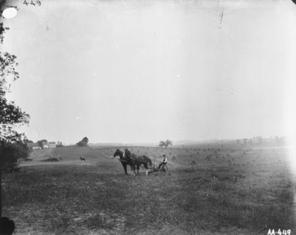 View across field towards man using a horse-drawn mower. In the background on the left is a person riding a horse near a large haystack, and behind the haystack are farm buildings. Sheaves of wheat and rolling hills are in the far distance.