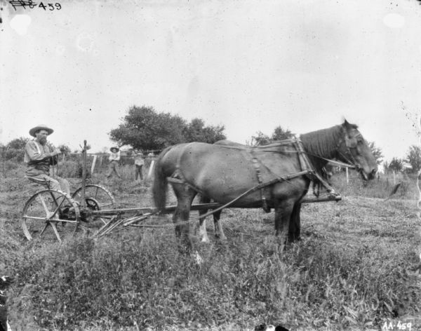 Right side view of a man sitting on horse-drawn mower in a field. In the background two young boys stand near a fence.