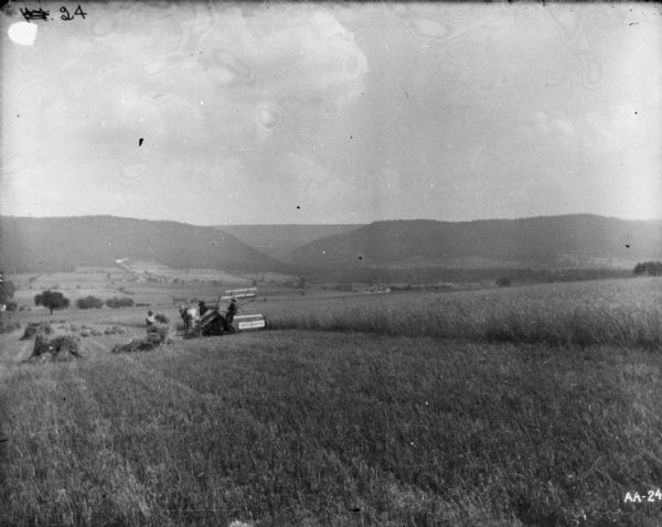 View down slope of hill towards a man standing in a field and working near a man who is using a horse-drawn binder in a field. A valley and hills are in the background.