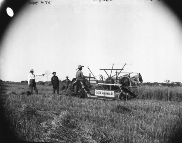 Men and boys are standing in a field,  near a man on a horse-drawn binder. One of the men is pointing to the right.
