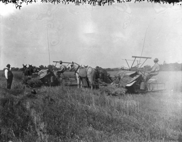 Two men are using horse-drawn binders in a field. A man wearing a vest and a hat is standing and watching on the left.