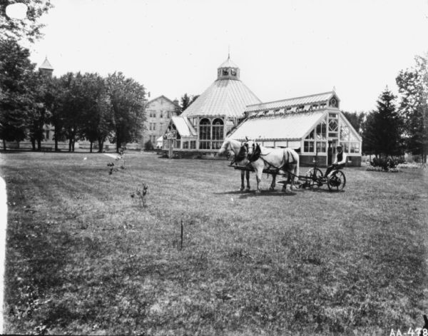 View across lawn of a man on a horse-drawn mower. In the background is a glass conservatory, and beyond that is a fence along a row of trees near a three-story stone building, and a tower rising above the treetops.