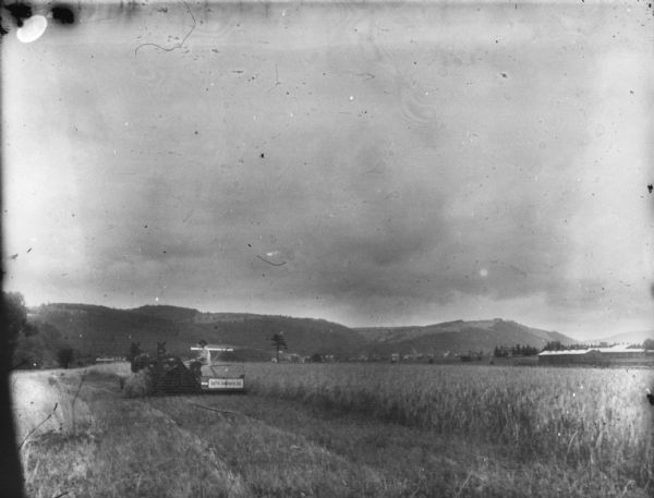 Rear view from a distance of a man using a horse-drawn McCormick binder in a field. There are buildings and hills in the far distance.