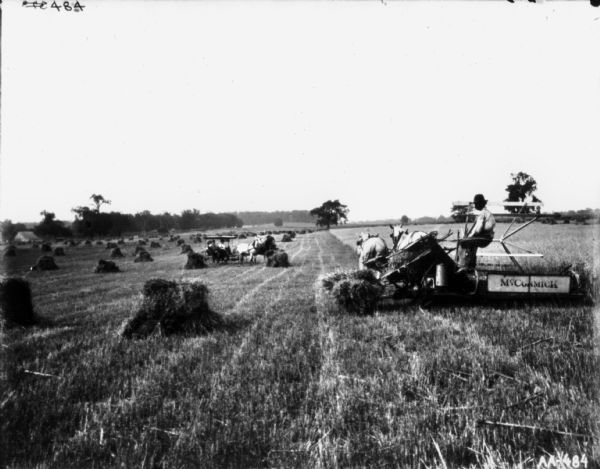 View towards a man using a horse-drawn McCormick binder in a field. Further down the field a man is driving a horse-drawn buggy. There is a farm building in the far background on the left.