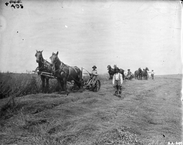 View towards two men in a field with horse-drawn binders. One man is standing in the foreground. In the background is a man standing next to another man in a horse-drawn cart.