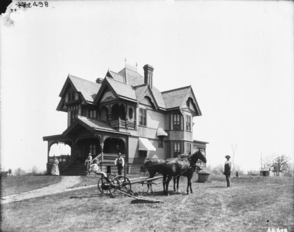 Group of people posing in front of large Victorian home (Joseph Robbins?). A child is sitting on a horse-drawn mower in the yard in front. The horses are wearing fly-nets. A woman, young boy, and man are posing in front of the porch. Another man wearing overalls is standing on the right. There is a balcony on the second floor of the house, and some of the windows have awnings.