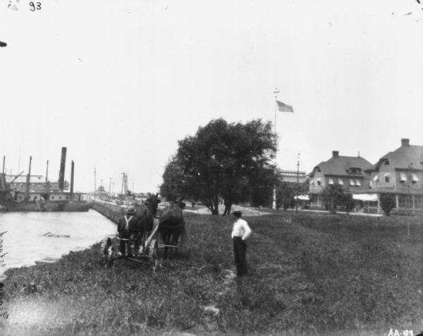 View from rear of a man using a horse-drawn mower along a shoreline at League Island. A man is standing near him on the right. In the background on the left are ships at dock, and buildings and trees are on the right.