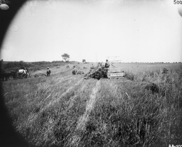View across field of harvested grain towards a man using a horse-drawn McCormick binder in a field. Two men are working with sheaves of wheat. Two other men are with a horse-drawn buggy on the far left at the edge of the field.