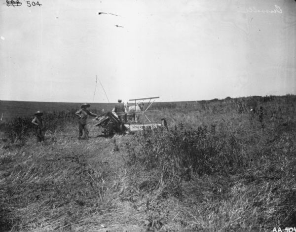 Rear view of a man using a horse-drawn McCormick binder in a field. A man is standing and leaning on the back of the binder, and a young man is standing on the left.