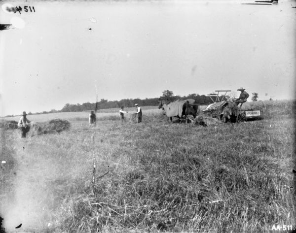 Four men working in a field near a man using a horse-drawn McCormick binder in a field. The horses are wearing blankets.