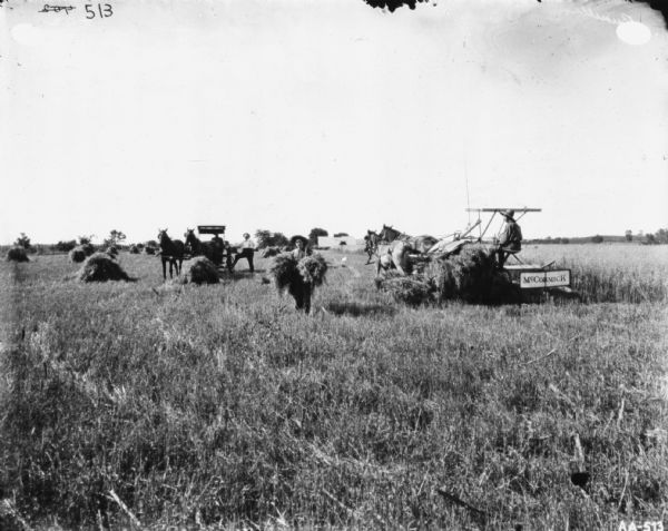 View towards men posing in a field with a horse-drawn McCormick binder. Two men are posing with a horse-drawn buggy in the center, and another man is standing holding two sheaves of wheat. A man on the right is using a horse-drawn binder. In the background are farm buildings.