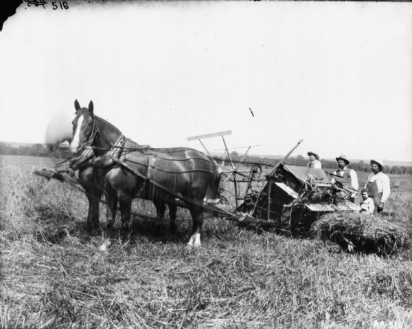 Two men and two children are posing with a horse-drawn binder in a field. The horses are wearing fly-nets.