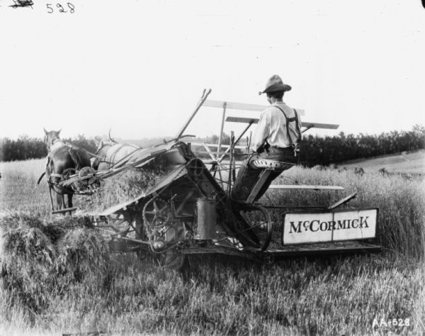 Three-quarter rear view from left of a man (D.R. Robinson?) using a horse-drawn McCormick binder in a field.
