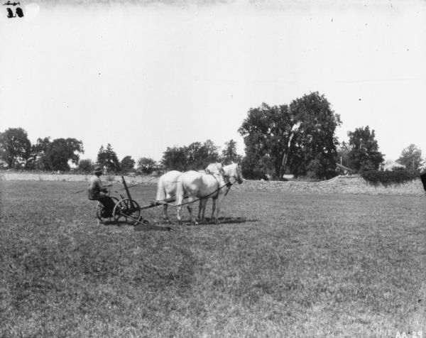 Man on horse-drawn McCormick mower in field. In the background is a stone wall. Behind the wall are trees and buildings.