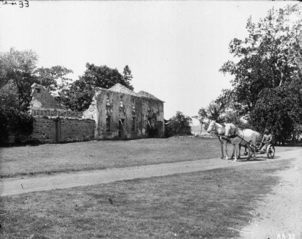 A man using a horse-drawn mower near the ruins of a stone building.