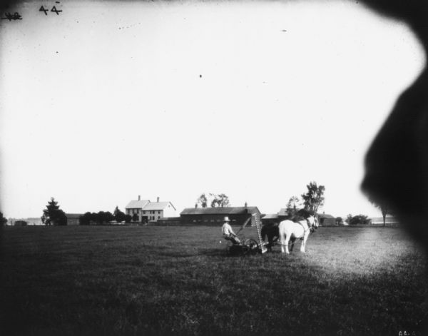 View across field of a man using a horse-drawn mower in a field. In the background are farm buildings.