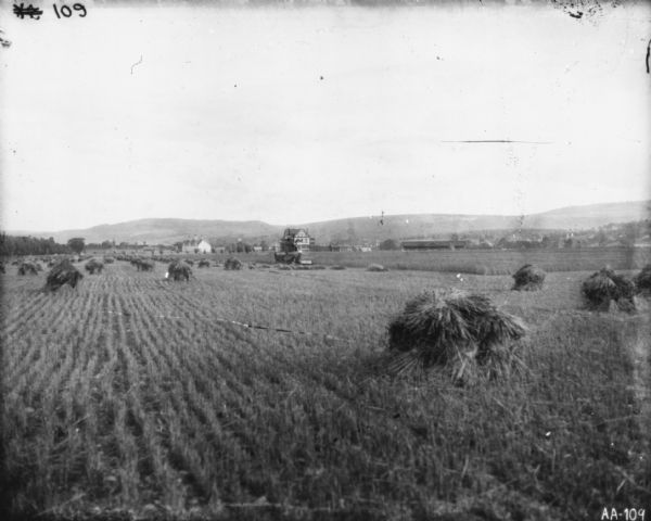 View across harvested field with sheaves of wheat towards a man using a horse-drawn McCormick binder in a field. In the far background are farm buildings and hills.