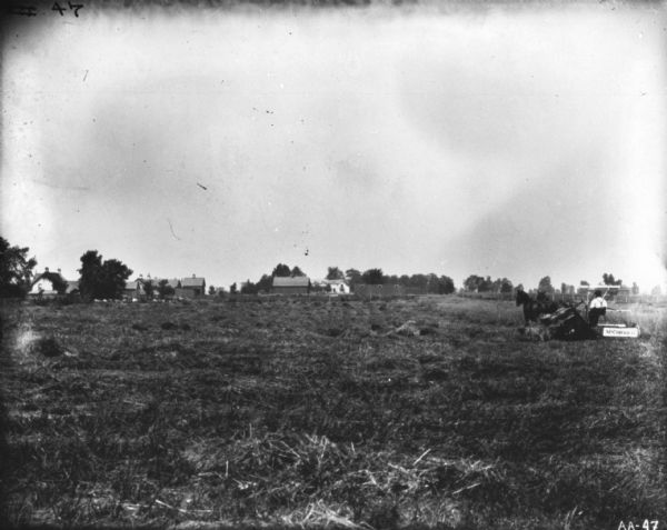 View across field of a man using a horse-drawn McCormick binder in a field. In the far background are farm buildings and a farmhouse.