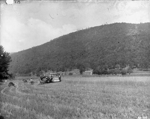 View across harvested field towards men and a young girl posing near a man using a horse-drawn McCormick binder. Farm buildings and fences are in the background at the base of a tall bluff or mountain.