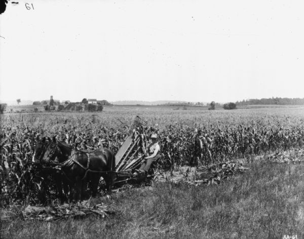 Left side view of a man using a horse-drawn corn binder at the edge of a field. In the far background are farm buildings.