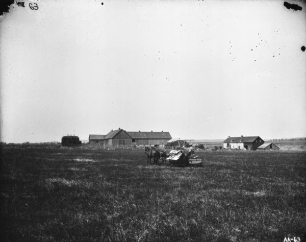 View across field of a man using a horse-drawn binder. In the background are farm buildings. Men are standing on a large stack of hay near barns on the left.