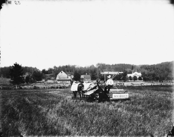 Rear view of a man using a horse-drawn binder in a field. There are farm buildings in the background, and trees and a low hill beyond.