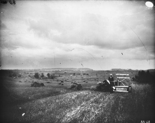 Rear view of a man using a horse-drawn McCormick binder in a field. Piles of sheaves are dotted throughout the hilly field. Hills and trees are in the far background.