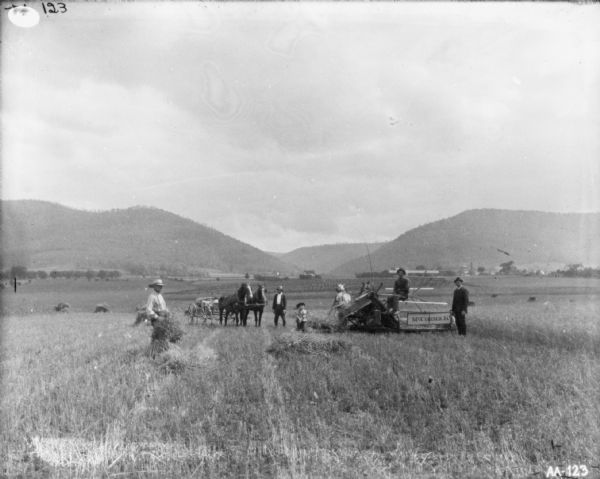 View across harvested field towards a group of people posing near a man in a horse-drawn McCormick binder. A man is standing on the left, and in the background a man is standing near a horse-drawn buggy. A young child is standing in the middle near the binder, and a man is standing on the right next to the binder. Behind them are farm buildings near the base of hills or mountains.