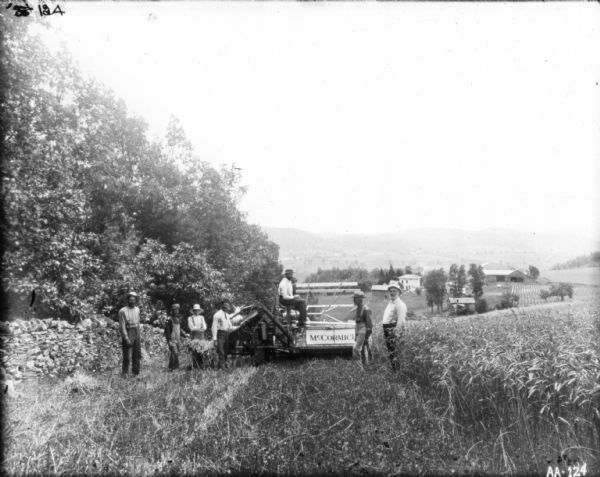 Group of men posing in field around a man sitting on a horse-drawn McCormick binder. In the background are farm buildings, and in the far distance a valley and hills.