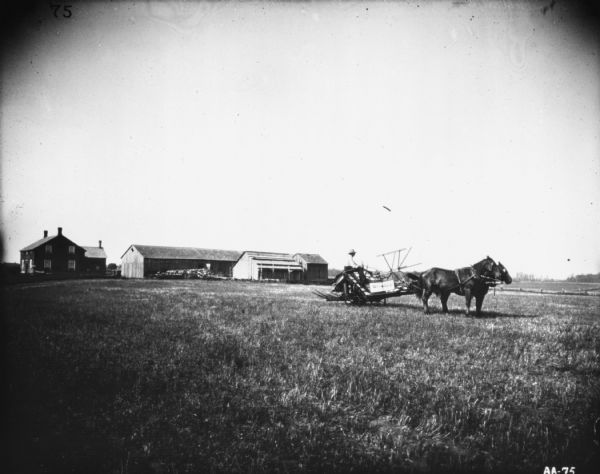 View of a man sitting on a horse-drawn McCormick binder on a transport truck in a field. There is a farmhouse and farm buildings in the background.