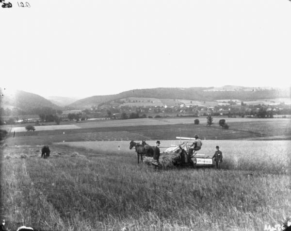 View across harvested field towards three men with a horse-drawn binder. The man on the left is wearing a suit and hat and is carrying a case. The man on the right us standing and holding the back of the McCormick binder. The other man is sitting on the binder. A horse is grazing on the far left. In the far background is a valley with hills beyond.
