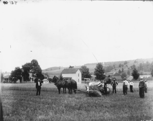 View across harvested field towards men posing near a man on a horse-drawn McCormick binder. Farm buildings are behind them, and in the far background are hills and trees.